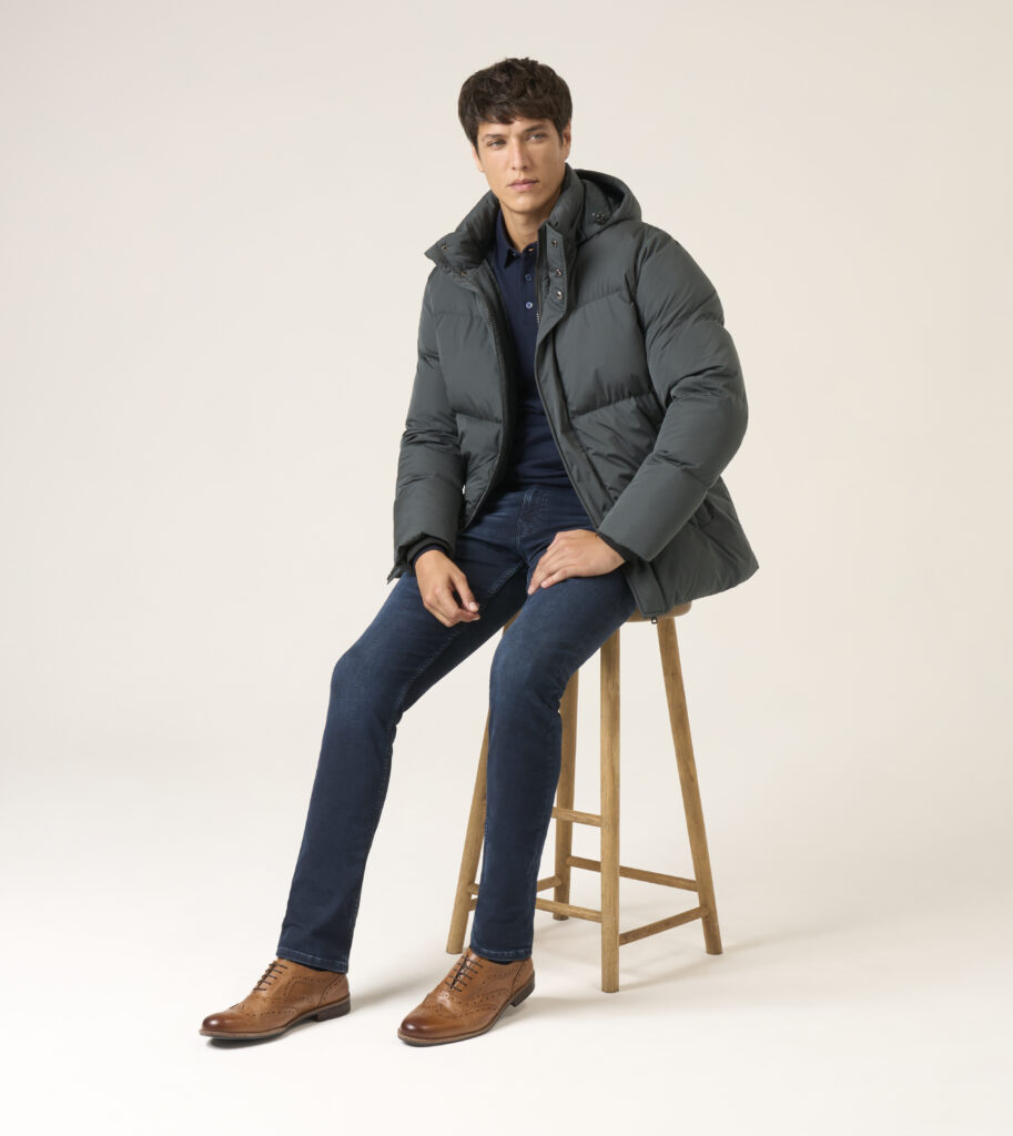 A man sitting on the stool. He is wearing navy sweater, puffer jacket, dark blue jeans and elegant brown shoes.